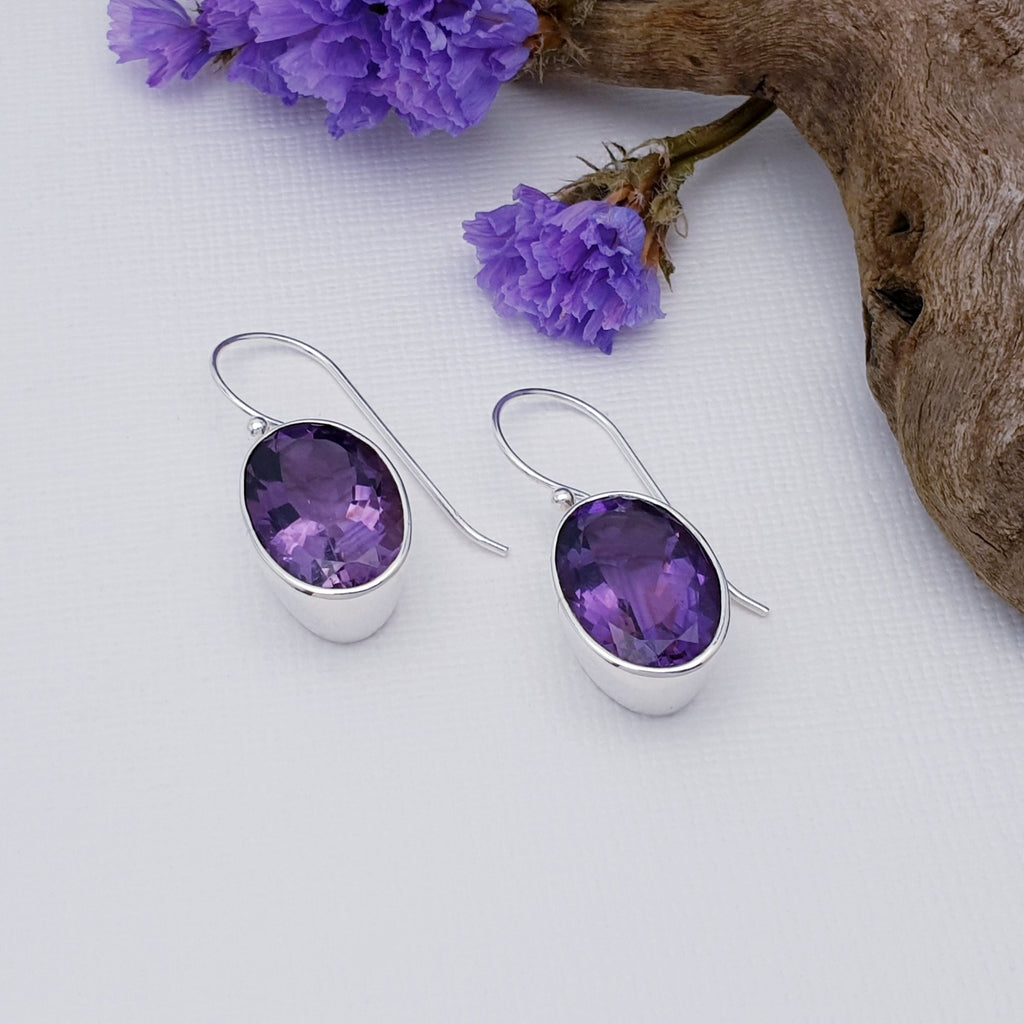 These earrings feature beautiful tabletop cut, oval, Amethyst stones in simple Sterling Silver settings. The Amethyst stones in these earrings are a beautiful deep purple colour, this is enhanced by the deep silver settings, creating a gorgeous and eye catching pair of earrings.