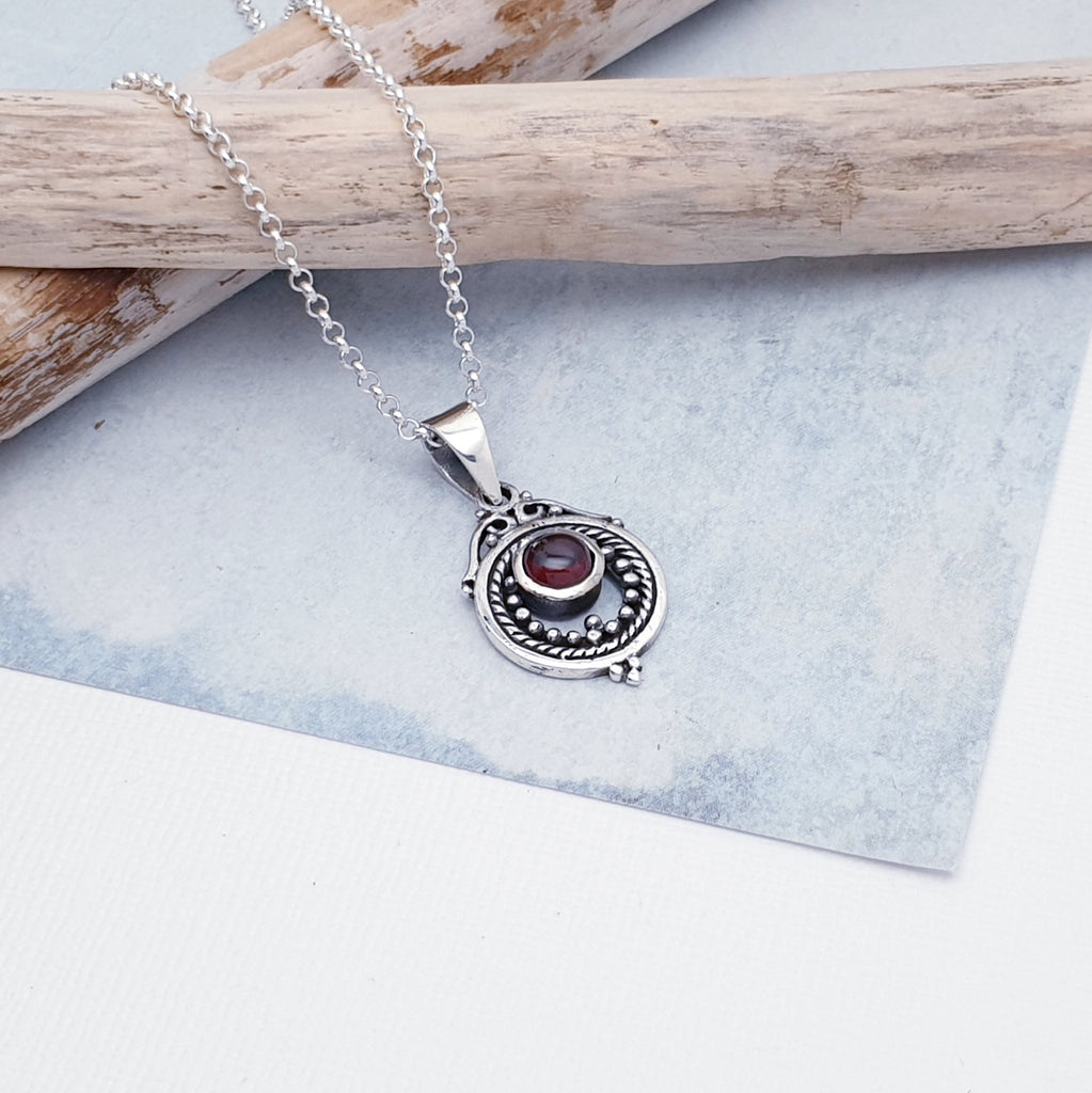 A gorgeous design, this pendant features a round, cabochon Garnet stone in a simple setting. Intricate Sterling Silver detailing decorates around the stone, and is given depth and presence by the clever use of oxidisation