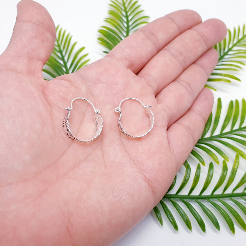 a pair of small sterling silver feather hoops in the palm of a hand
