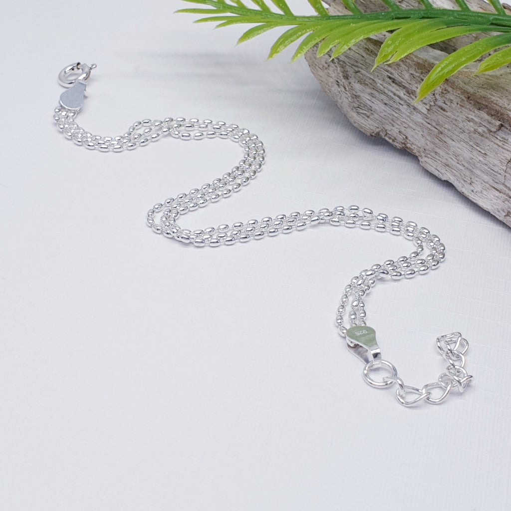 Delicate, adjustable sterling silver bracelet with three strands of tiny oval beads on a fine chain, displayed on a white background with wood and foliage as decorations