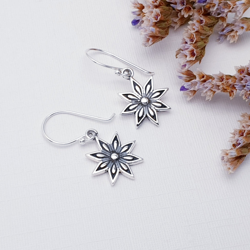 Silver star dangle earrings with a detailed middle, on a white background with multi coloured flowers in the corner.