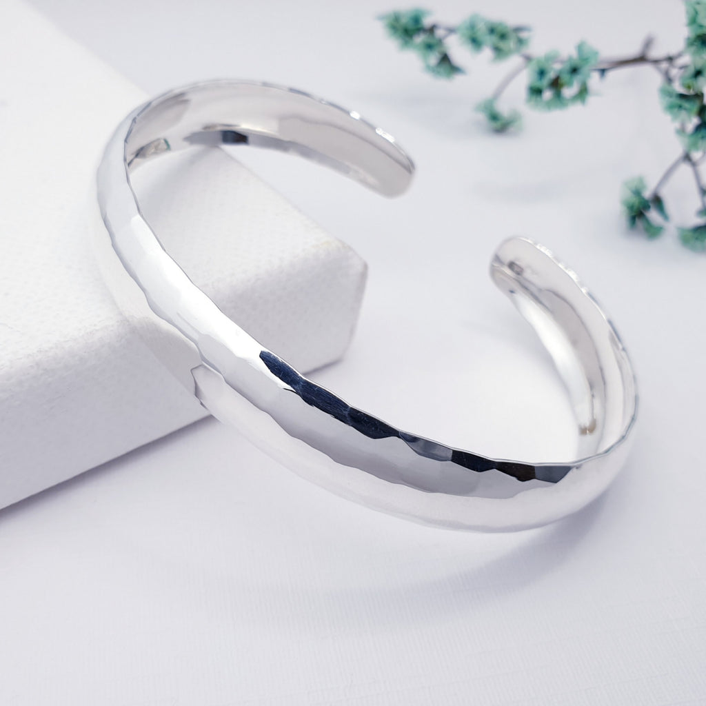 Sterling silver hammered cuff bracelet on a white background with green foliage in the top right corner of the image.