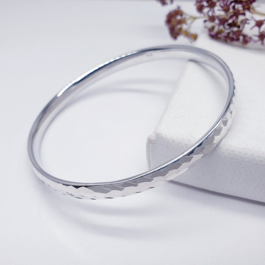 Sterling silver checkerboard bangle on a white background with purple foliage in the top right hand corner.