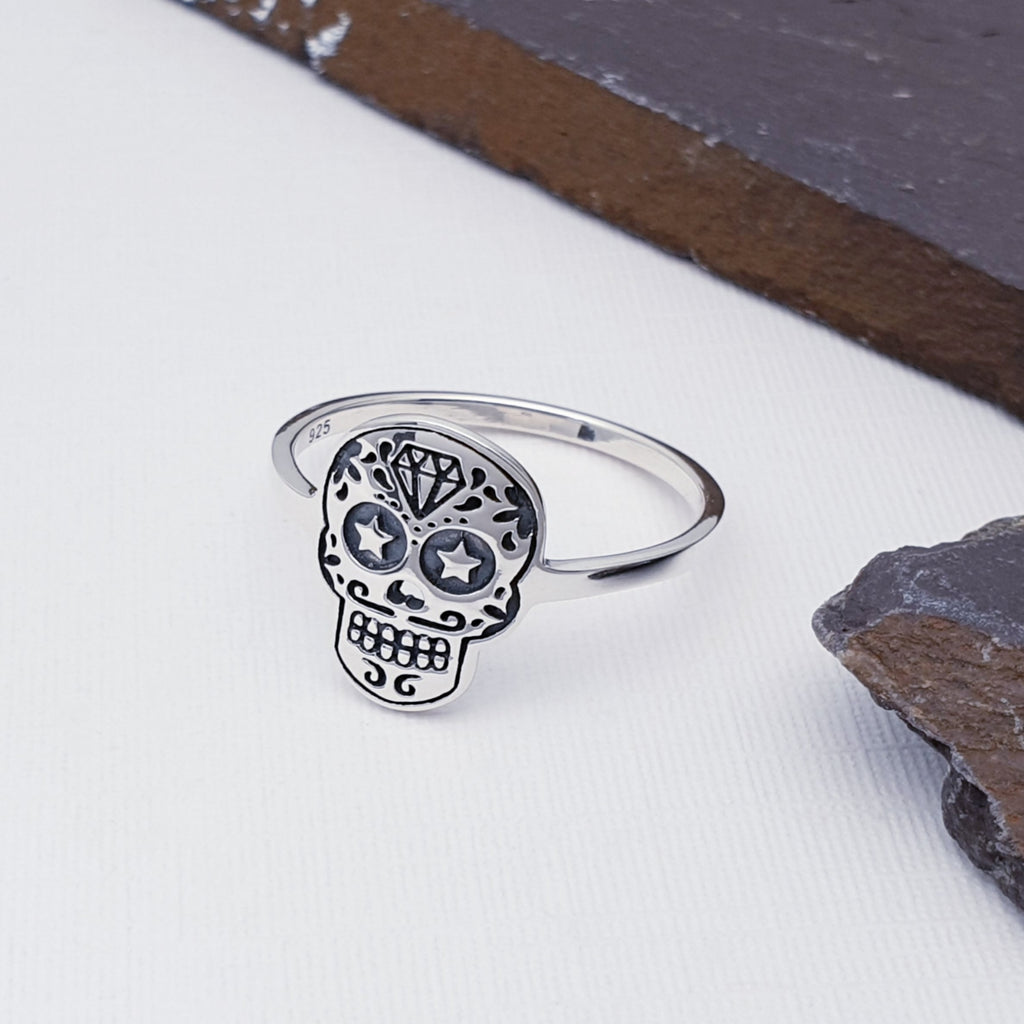 Sterling silver sugar skull ring on a white background with slate around the right edge of the image. The sugar skull itself features a diamond on the forehead, stars in the eye sockets and a fun goatee around two rows of teeth.