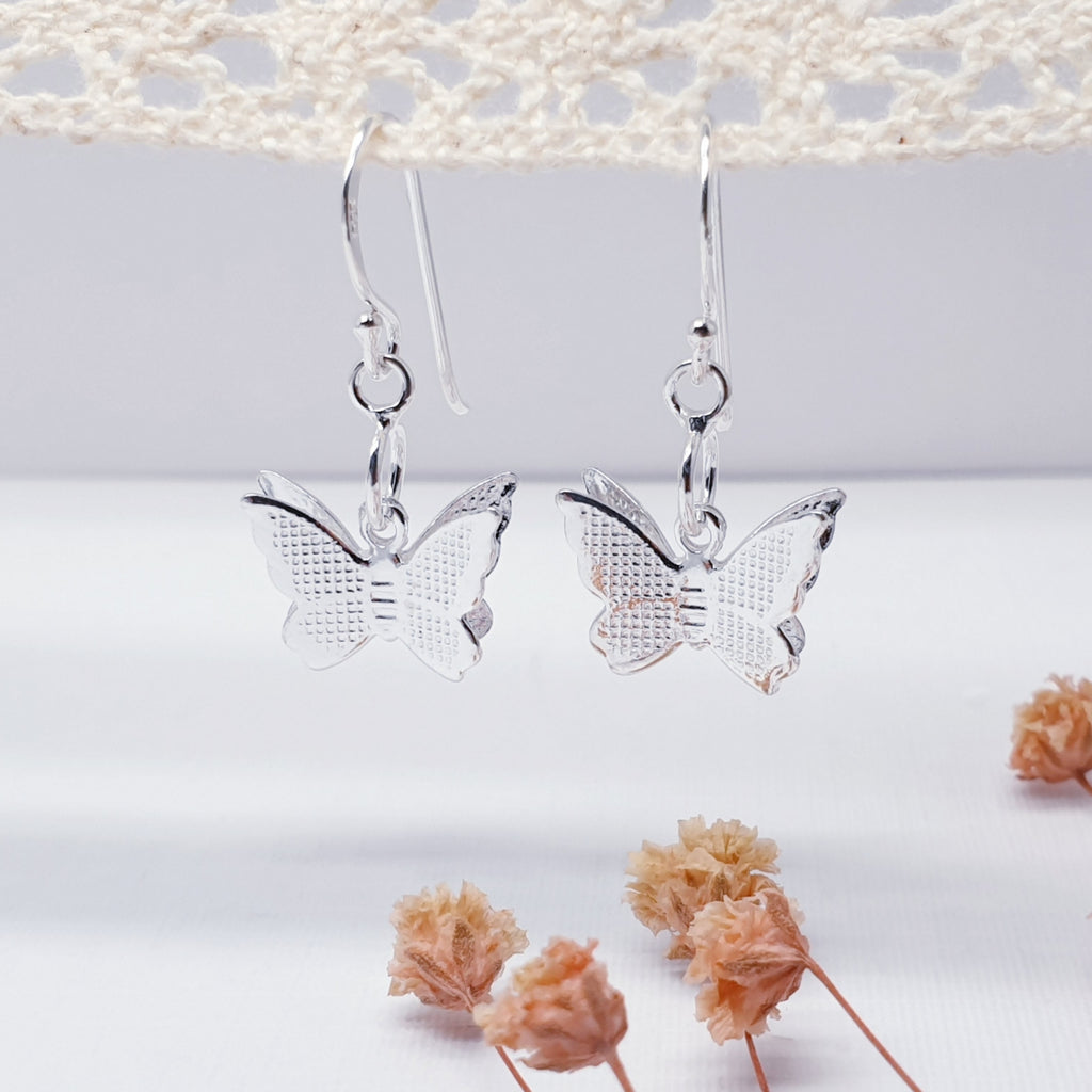 Sterling Silver Double Butterfly Earrings looped onto a piece of crochet with flowers on the bottom portion of the background. The butterflies are in the centre of the image