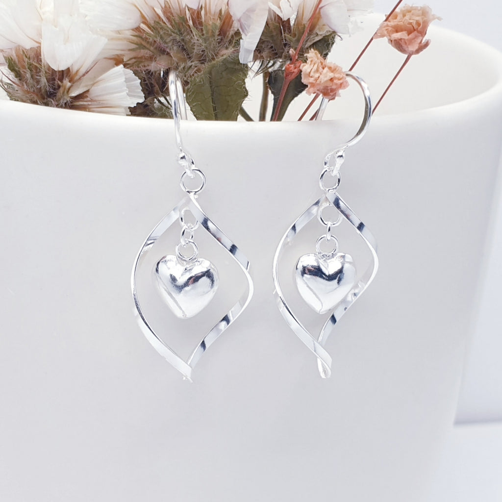 Sterling Silver Twisted Heart Earrings hanging from a plant pot which has pink and white flowers inside, along with leaves