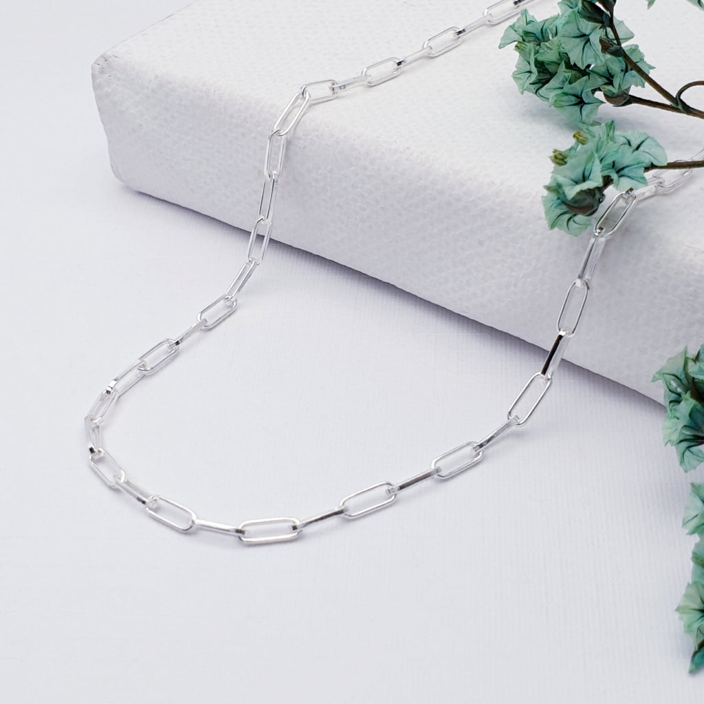 Our Sterling Silver paperclip chain displayed on a white background with teal coloured flowers in the background as decorations