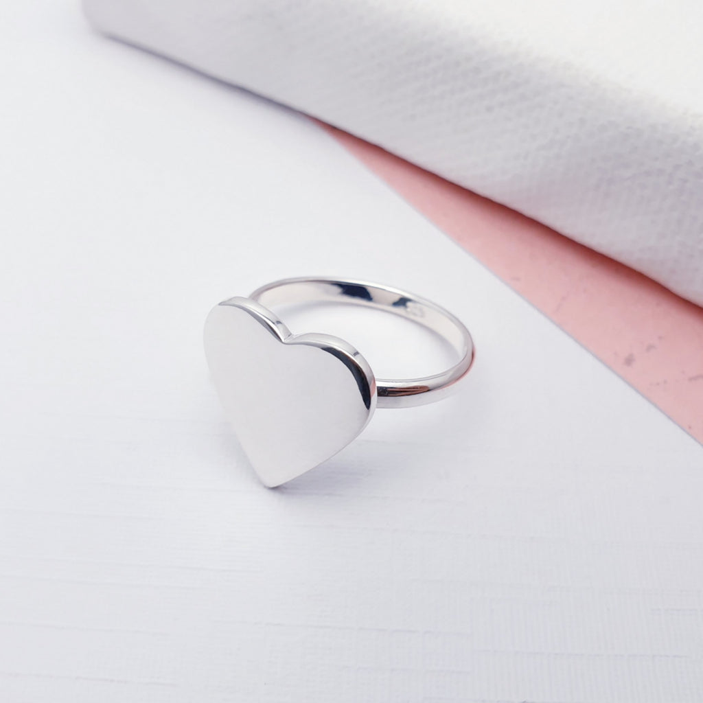 Our Sterling Silver polished heart ring displayed on a white background with pink card in the background as decoration