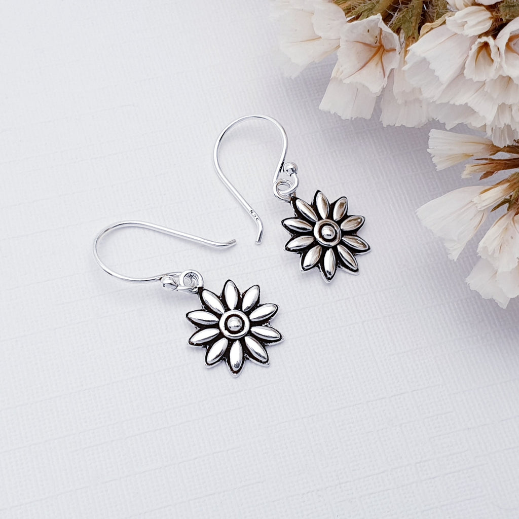 Our Sterling Silver Daisy earrings displayed against a white background with white flowers in the background as decoration
