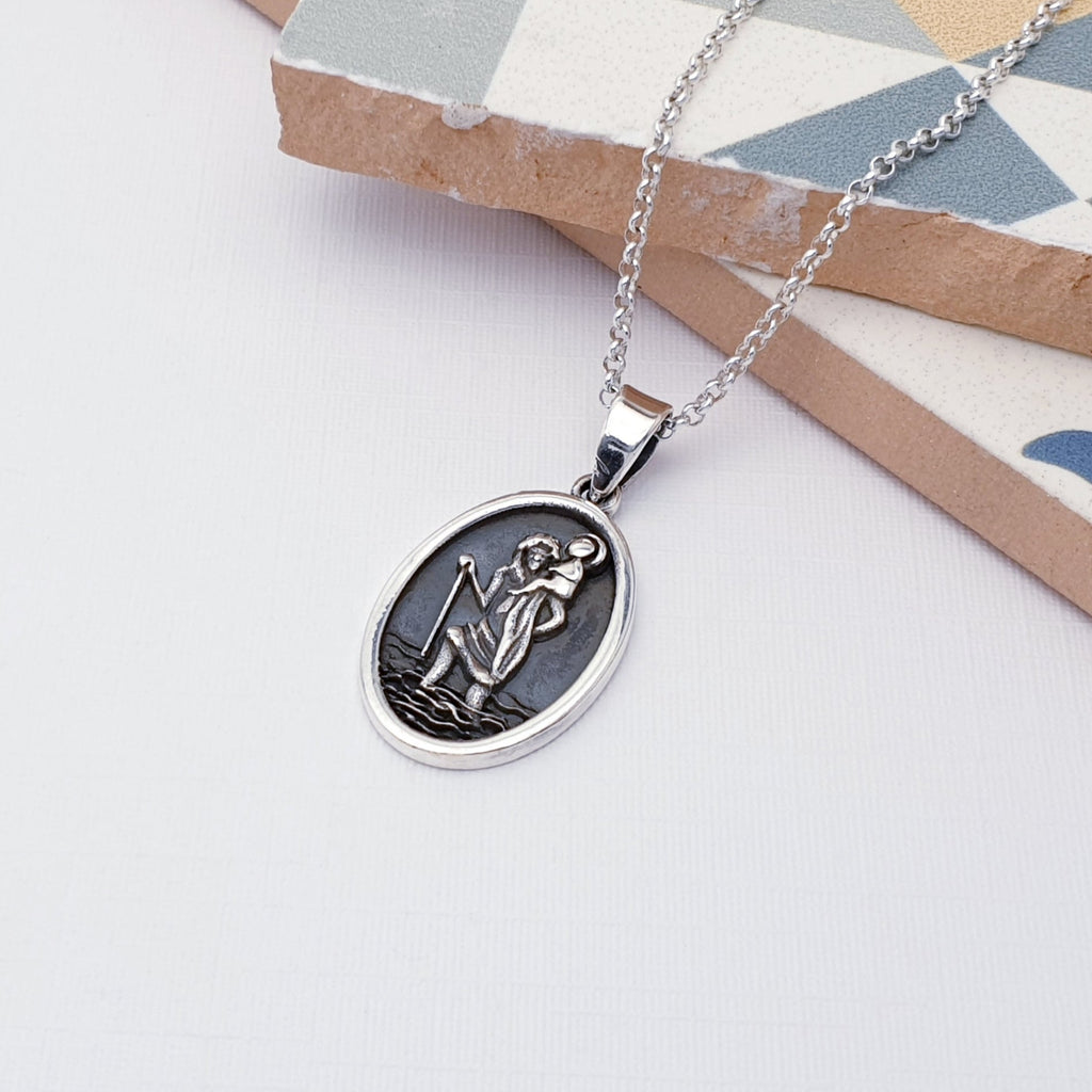 Our oval sterling silver st christopher pendant on a fine belcher chain, draped across some mosaic tiles and displayed on a white background.