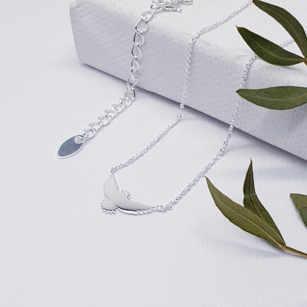 Our Sterling Silver flying bird necklace displayed a white background and draped over a white canvas with green leaves on one side as decoration.