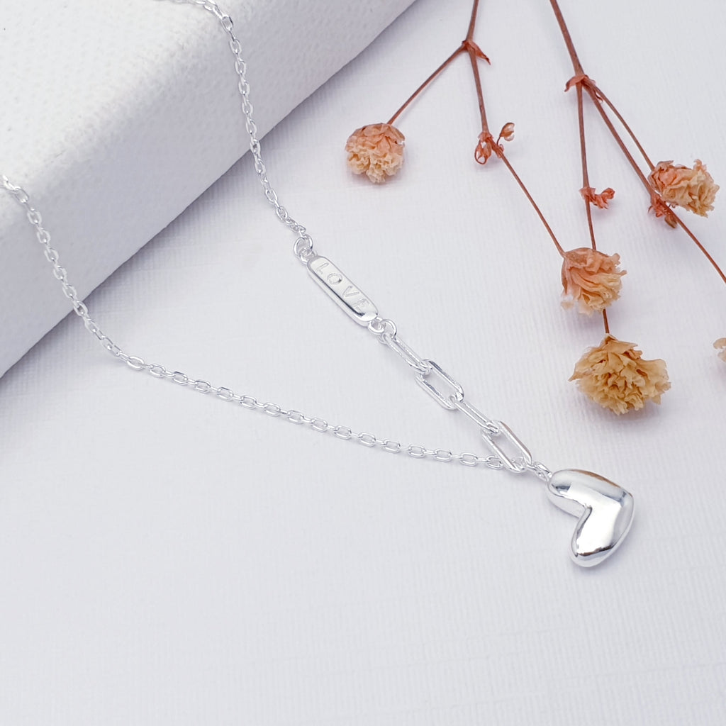 Our sterling silver love necklace displayed on a white background, draped over a white canvas, with pink flowers on the right as decoration