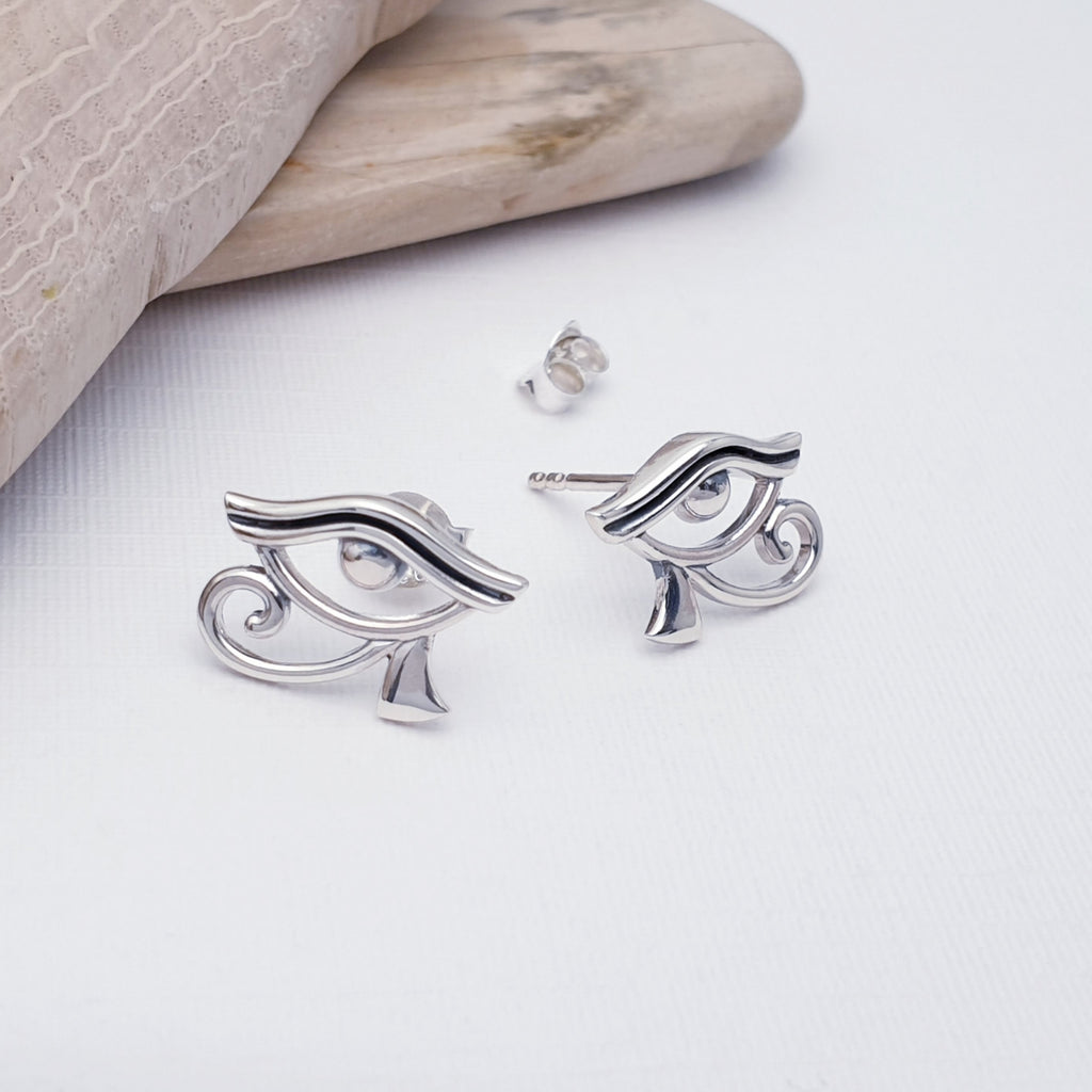 Silver eye-shaped earrings on a light surface with a wooden block in the background