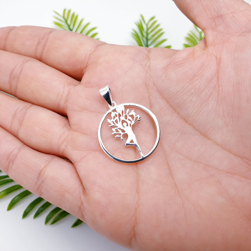 Silver tree pendant held in a hand with green leaves in the background