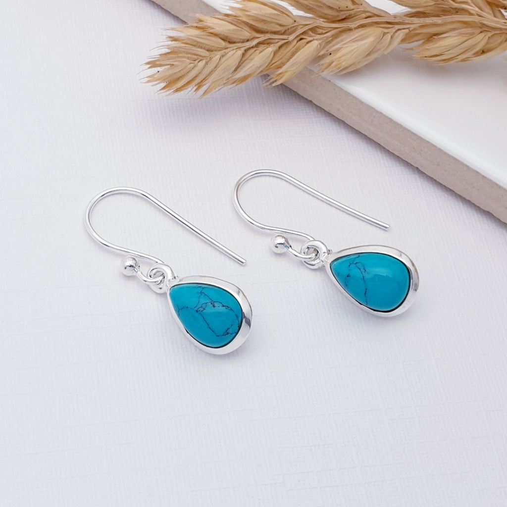 Our simple turquoise teardrop earrings displayed on  a white background with a white tile and autumn foliage decorating the back of the picture