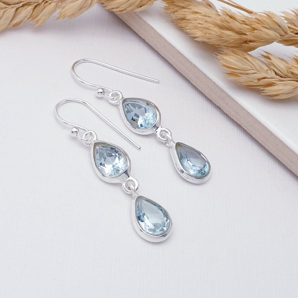 Double teardrop blue topaz earrings displayed on a white background with autumnal foliage decorating the background