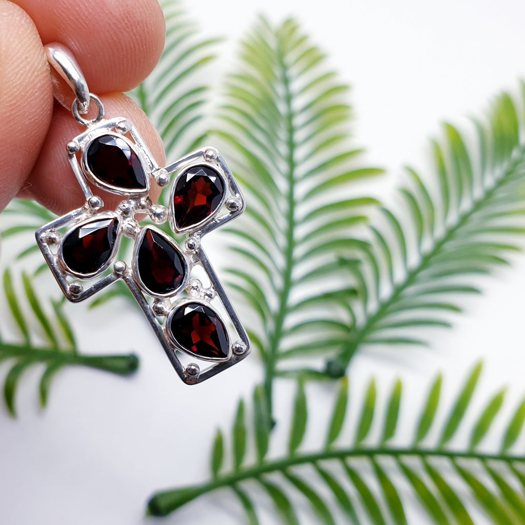 sterling silver cross pendant with garnet teardrop gemstones with foliage behind.