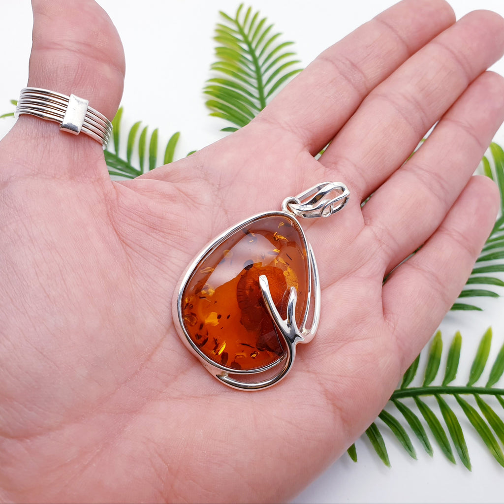 A large toffee amber pendant, with natural inclusions, set in sterling silver with branch detailing. In the palm of a hand with green foliage in the background