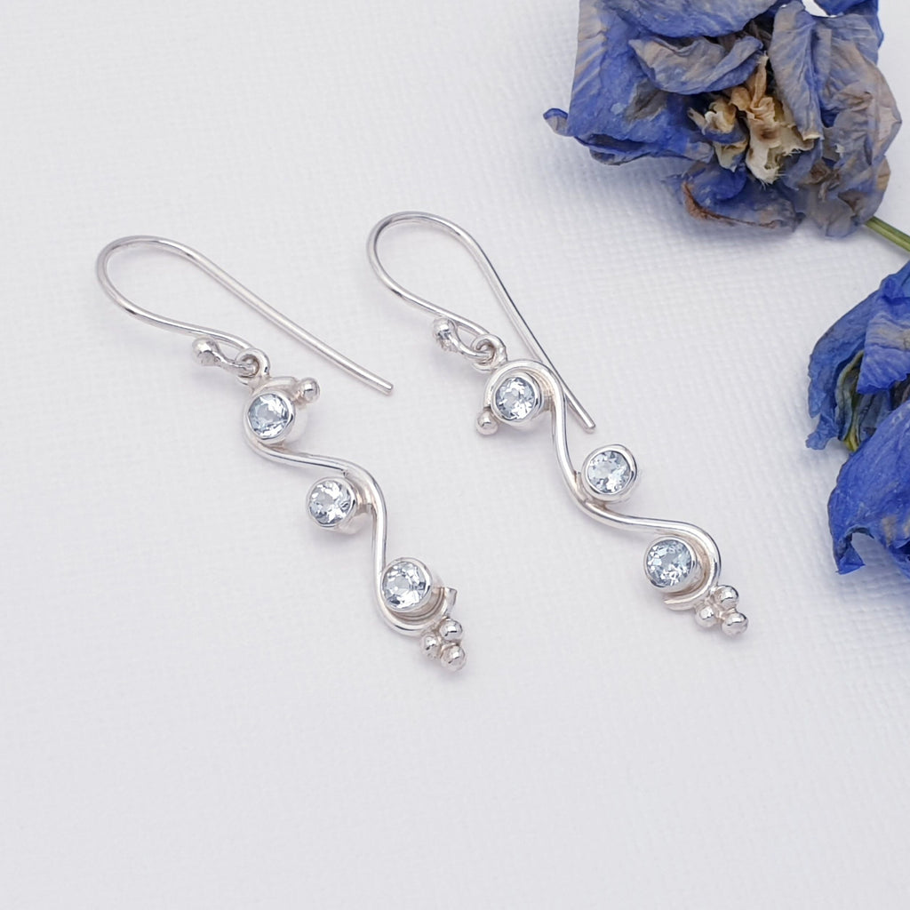 blue topaz gemstone and sterling silver swirly drop earrings flat lay with blue petals