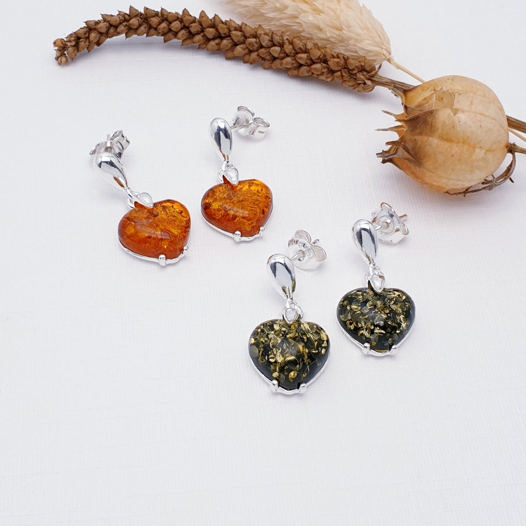 two pairs of earrings in toffee amber and green amber. The stones are heart shaped and dangle from a teardrop stud attachment. The earrings are layed flat on a white background with autumn foliage decorations.