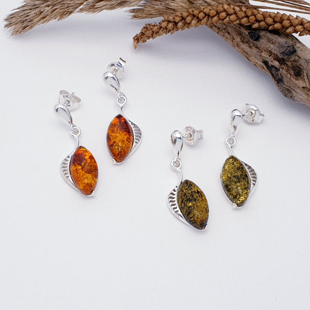 Tow pairs of earring in Toffee and Green Amber. The stones are marquise shaped in simple settings. the settings widen on one side with delicate cut out line shapes giving them a fish fin or fish scales feel. The earrings are layed flat on a white background with autumn leaves and wood decorations