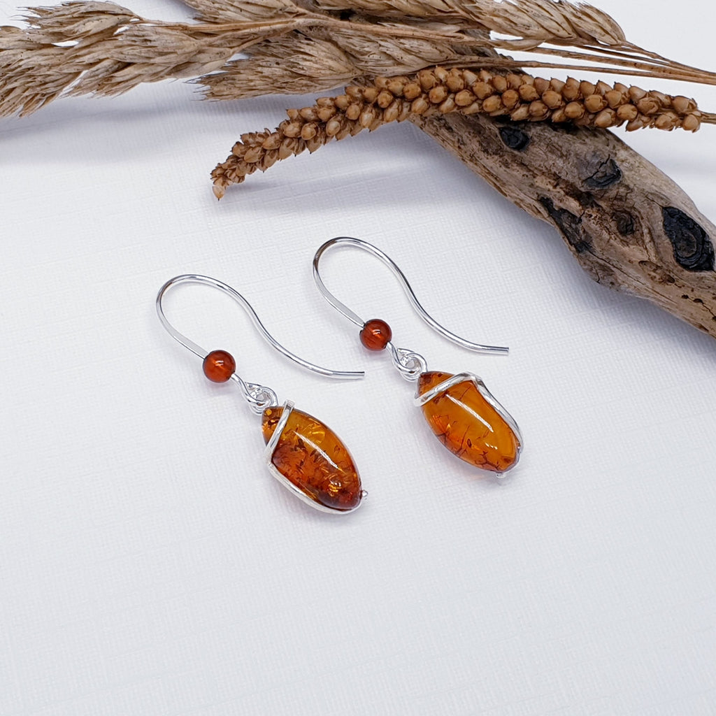 A pair of our Amber Gisela earrings, featuring oval stones with silver wire wrapped round. A small amber bead decorates the bottom of the earring hook. The earrings are layed flat on a white background with wood and autumn foliage as decorations.