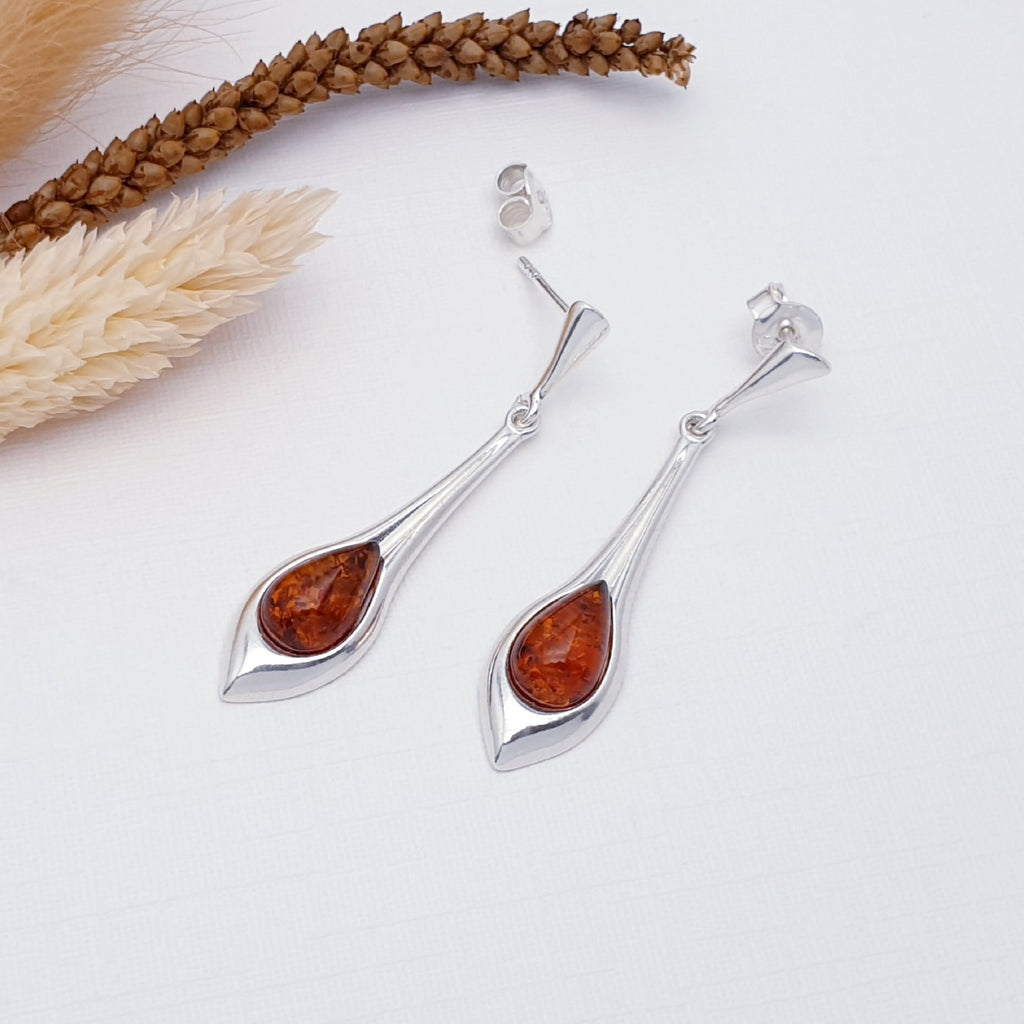 A pair of our toffee amber ozella earrings, featuring teardrop amber stones in a long fluid tear drop setting, with triangular stud attachments. They are layed flat on a white background with autumn foliage decorations.