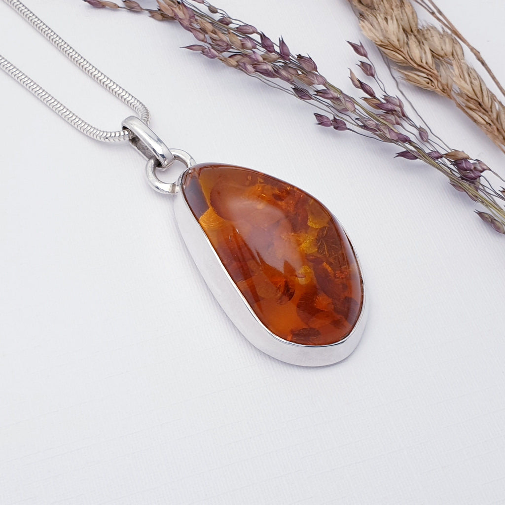 Our Toffee Amber Kasia Pendant on a heavy Snake chain, displayed on a white textured background with foliage decorations