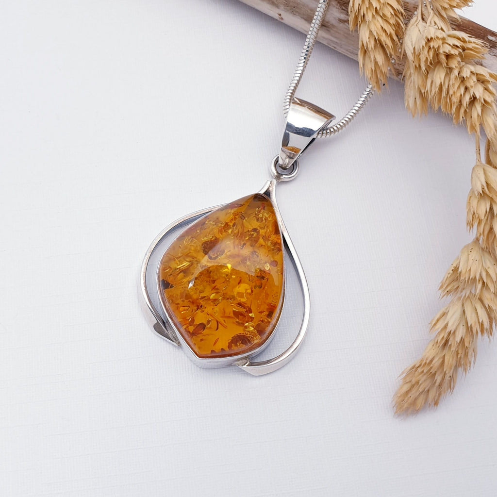 Our Baltic Toffee Amber Malgosia pendant on a Heavy Snake chain, displayed flat against a white background, with wood and foliage decorations.