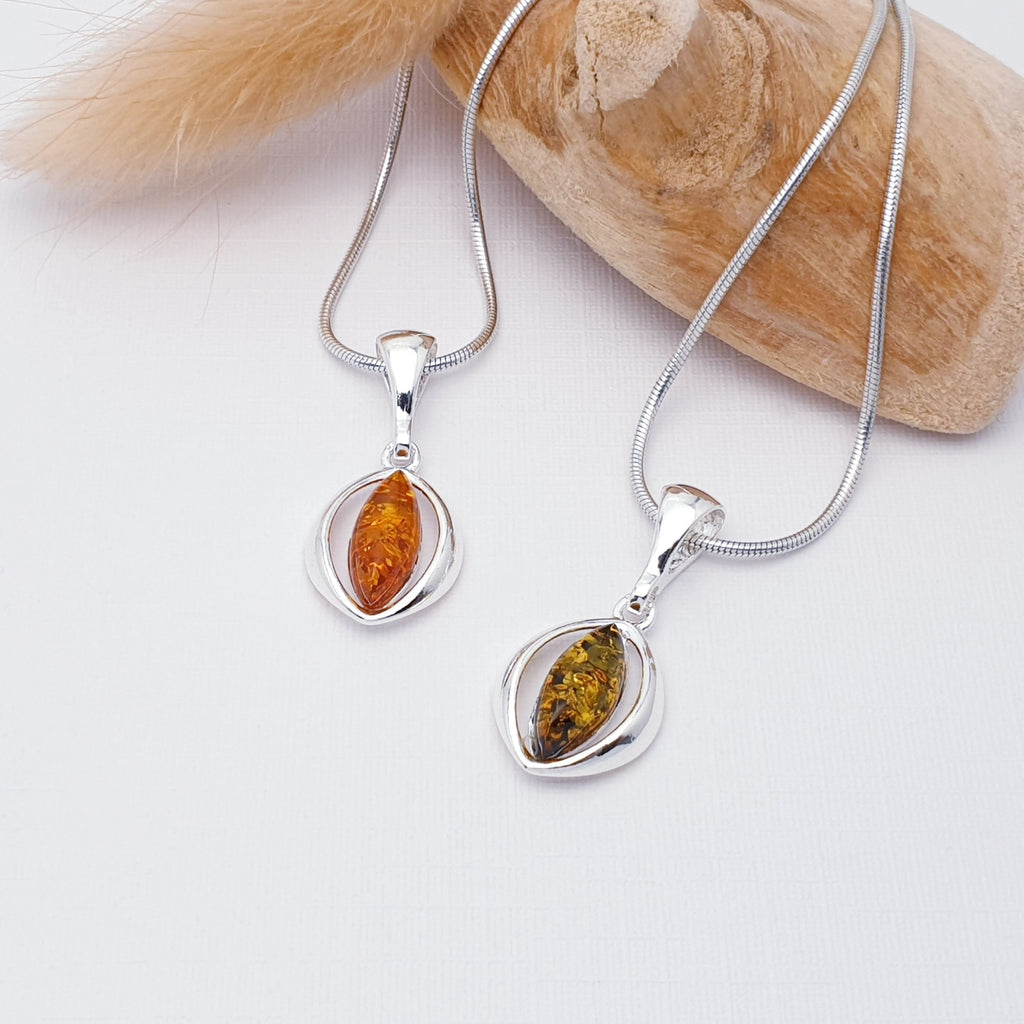 Two of our amber seed pendants, one toffee amber and one green amber, both strung on fine snake chains. The pendants are layed flat on a white background with drift wood and autumn foliage as decorations.