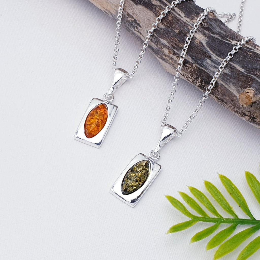 Two Rectangle shaped amber pendants with a marquise shaped amber at its center, one in toffee amber and one in green amber. These are strung on fine belcher chains. The pendants are layed flat against a white background with wood and a green leaf as decorations