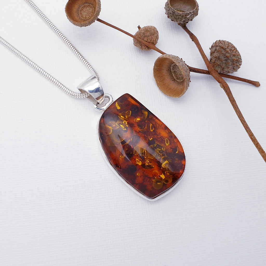 Our Dark Toffee Amber Loda pendant strung on a heavy snake chain, layed flat on a white background with seed pods decorations