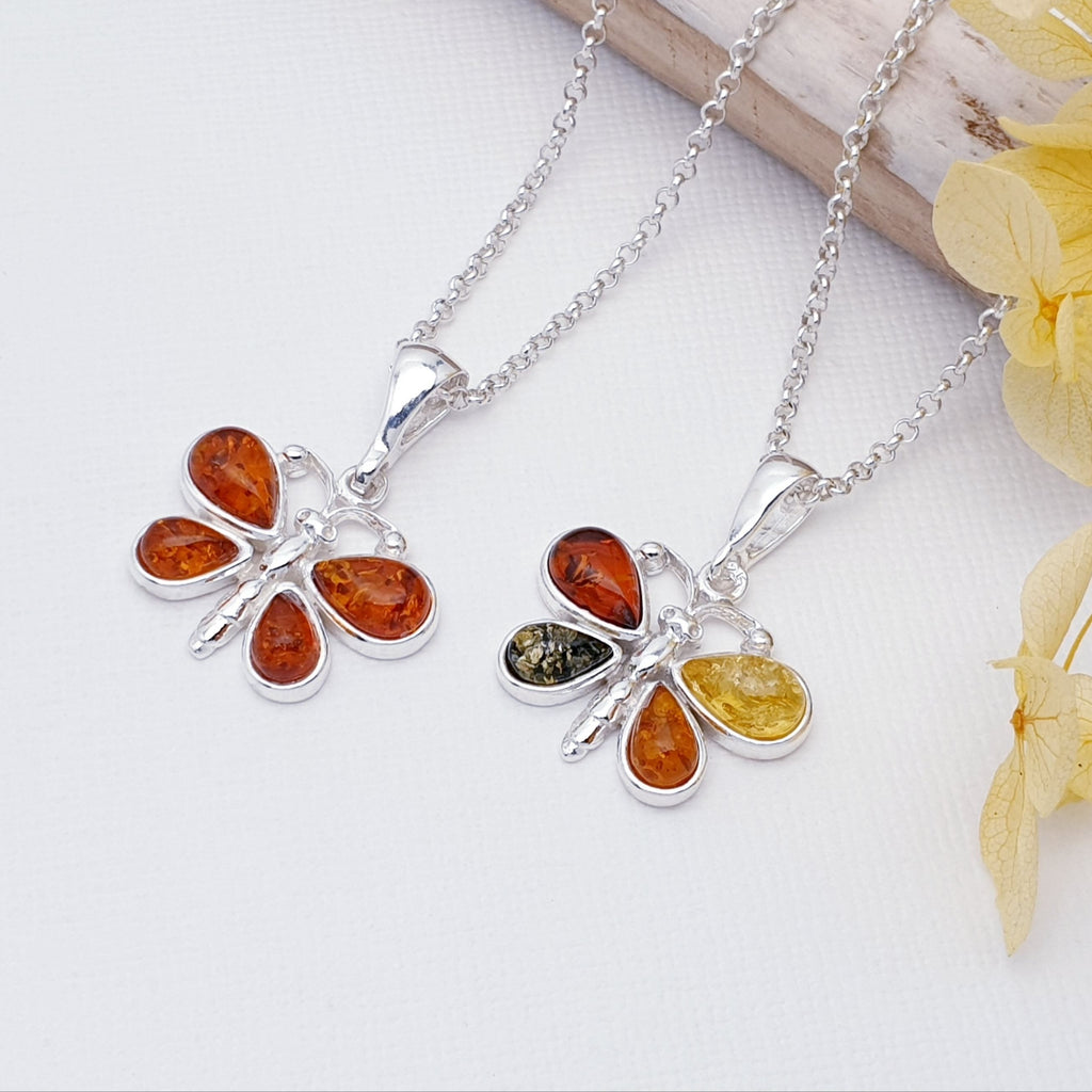 Two Amber butterfly pendants, one in toffee amber and the other in mixed amber, strung on fine belcher chains. Layed flat against a white background with wood and yellow flowers as decorations