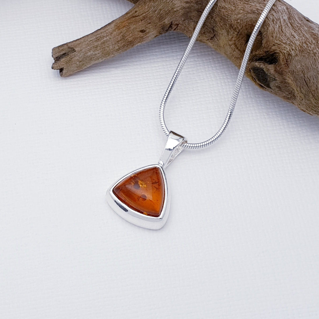 Our Toffee Amber simple triangle pendant strung on a fine snake chain, layed flat on a white background with driftwood decorations