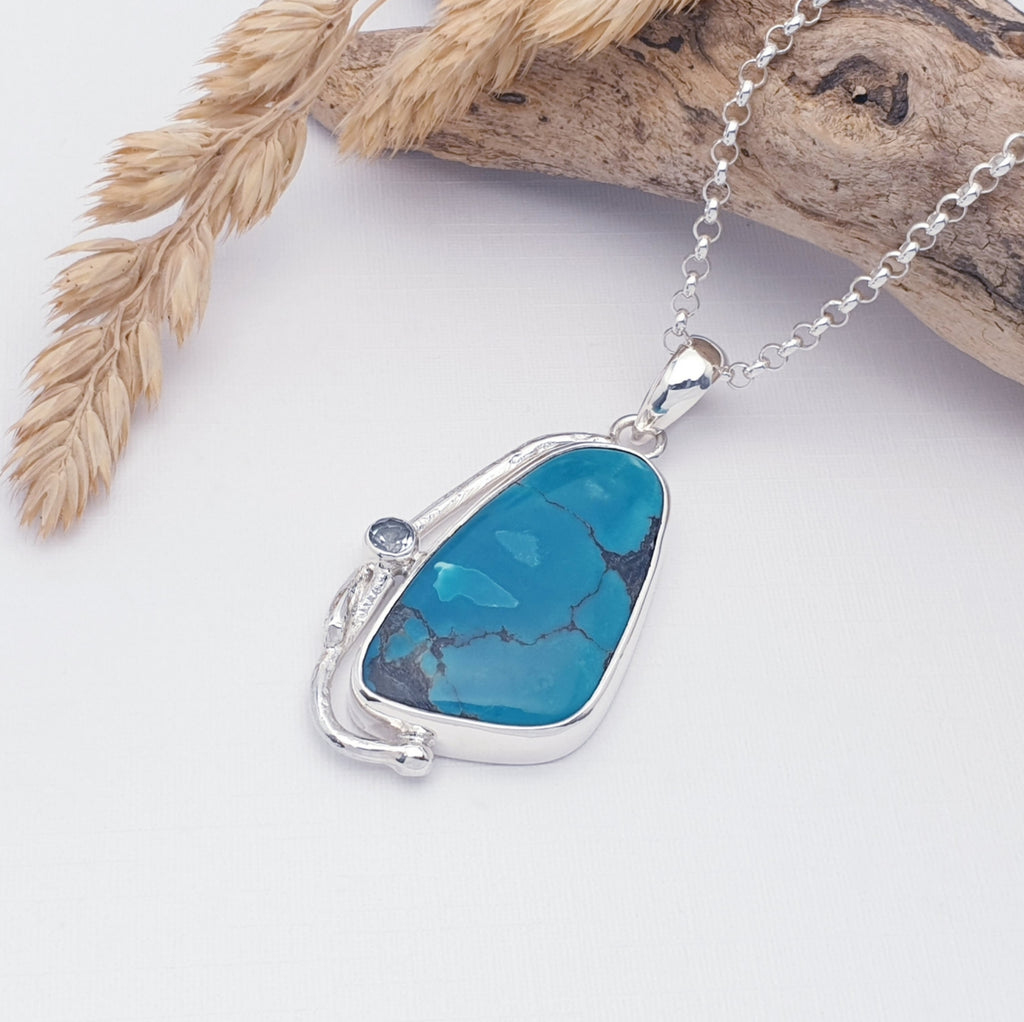 turquoise and blue topaz gemstone pendant with sterling silver botanical design, displayed on a white background with driftwood and autumnal foliage as decorations