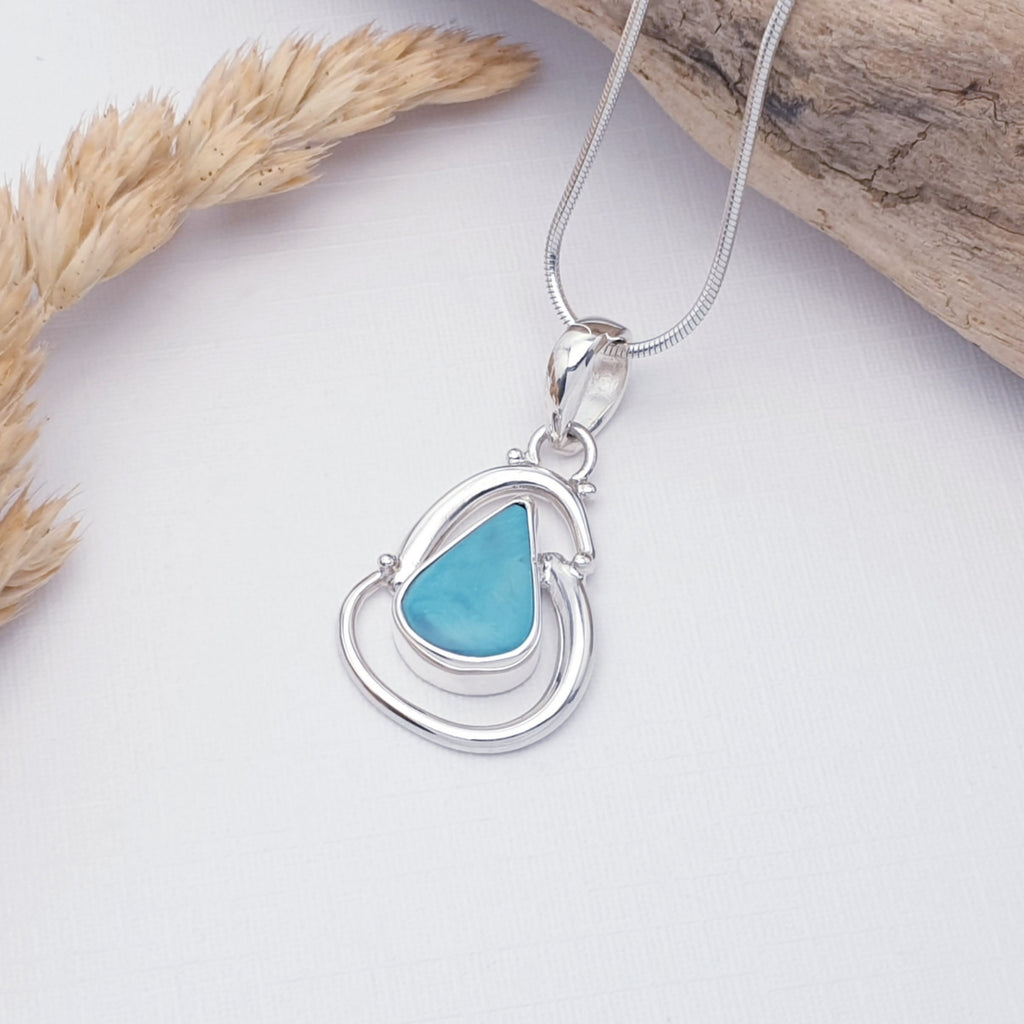 Handmade turquoise gemstone pendant with swirling sterling silver design surrounding the stone, displayed on a white background with driftwood and autumnal foliage as decorations
