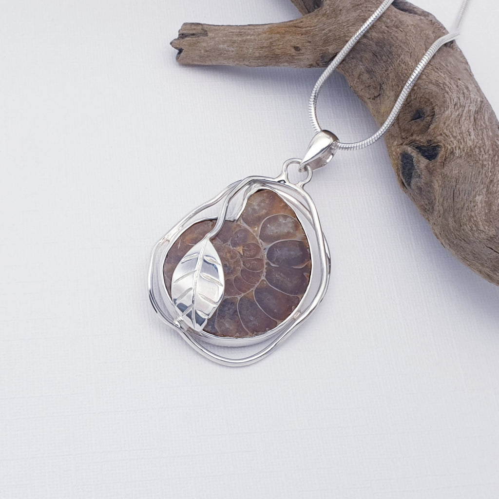 Ammonite fossil pendant with sterling silver leaf design, displayed on a white background with driftwood as decoration