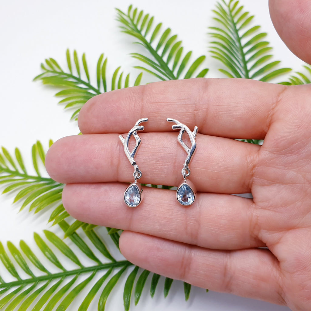 Our blue topaz vine earrings pictured on a hand to give impression of size, with green leaves in the background