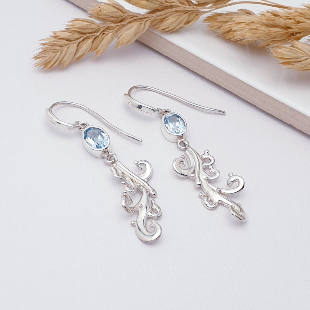 Our blue topaz sea breeze earrings displayed on a white background with a white tile and foliage as decorations