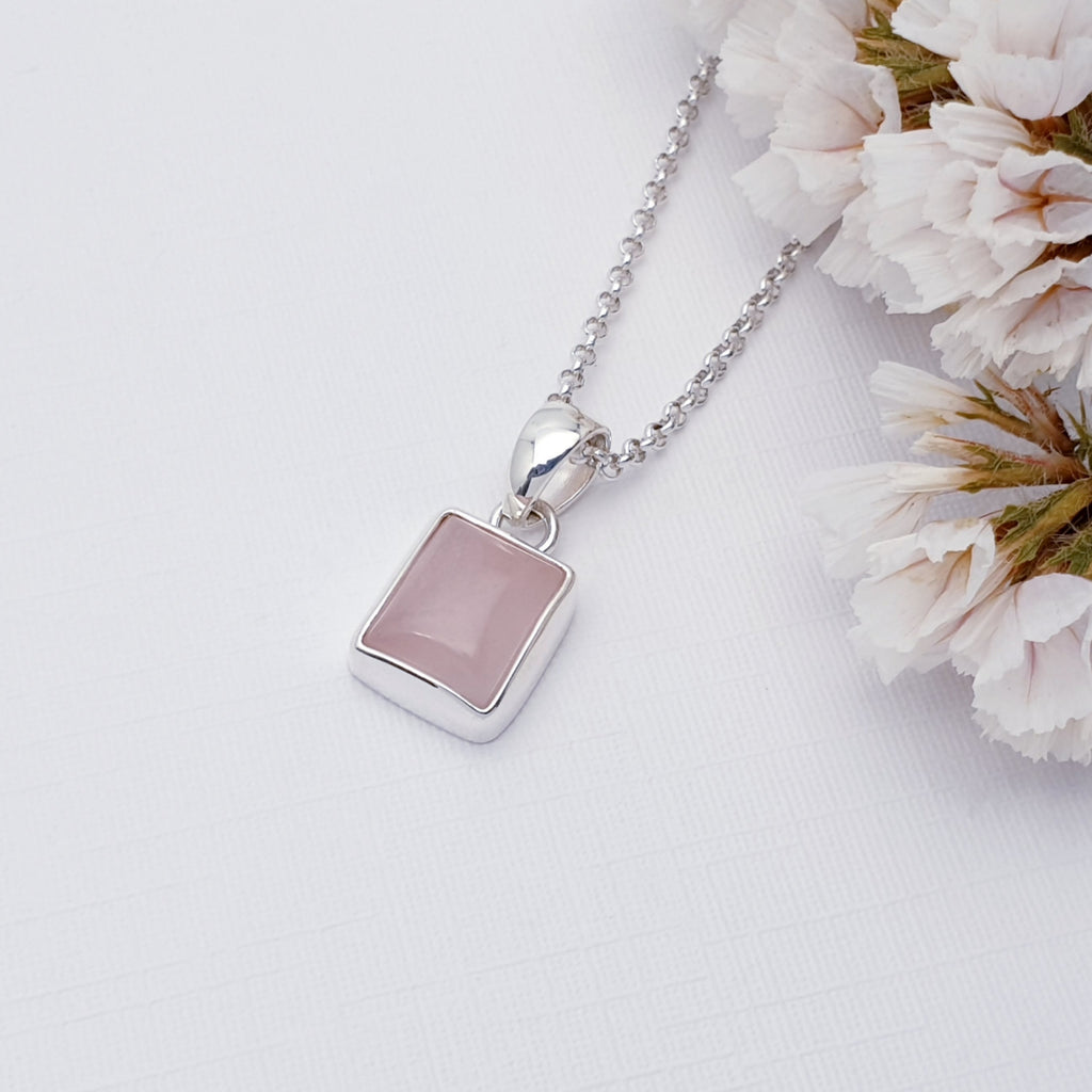 Our Rose Quartz Sterling Silver Rectangle pendant with a fine belcher chain, photographed against a white background with white flowers as decoration on the right had side of the picture