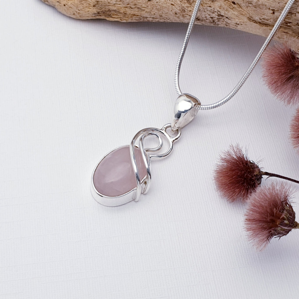 Our Rose Quartz Sterking Silver Cora pendant on a fine snake chain, photographed against a white background with wood and maroon flowers as decorations.