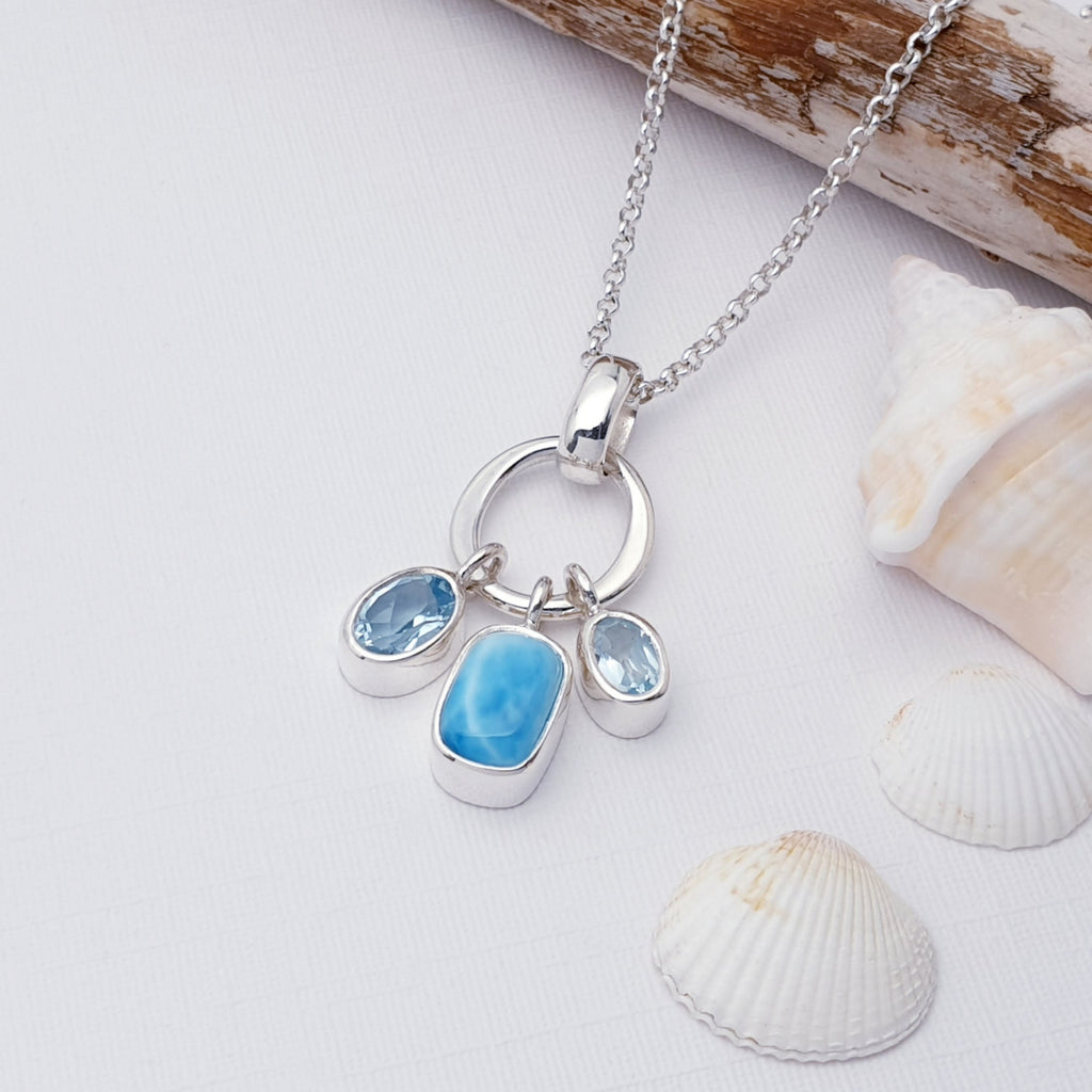 Blue Topaz and Larimar Pendant. Larimar in the centre in a rectangle cut and Blue Topaz in two oval cuts. It is displayed on a white background with sea shells and drift wood in the foreground