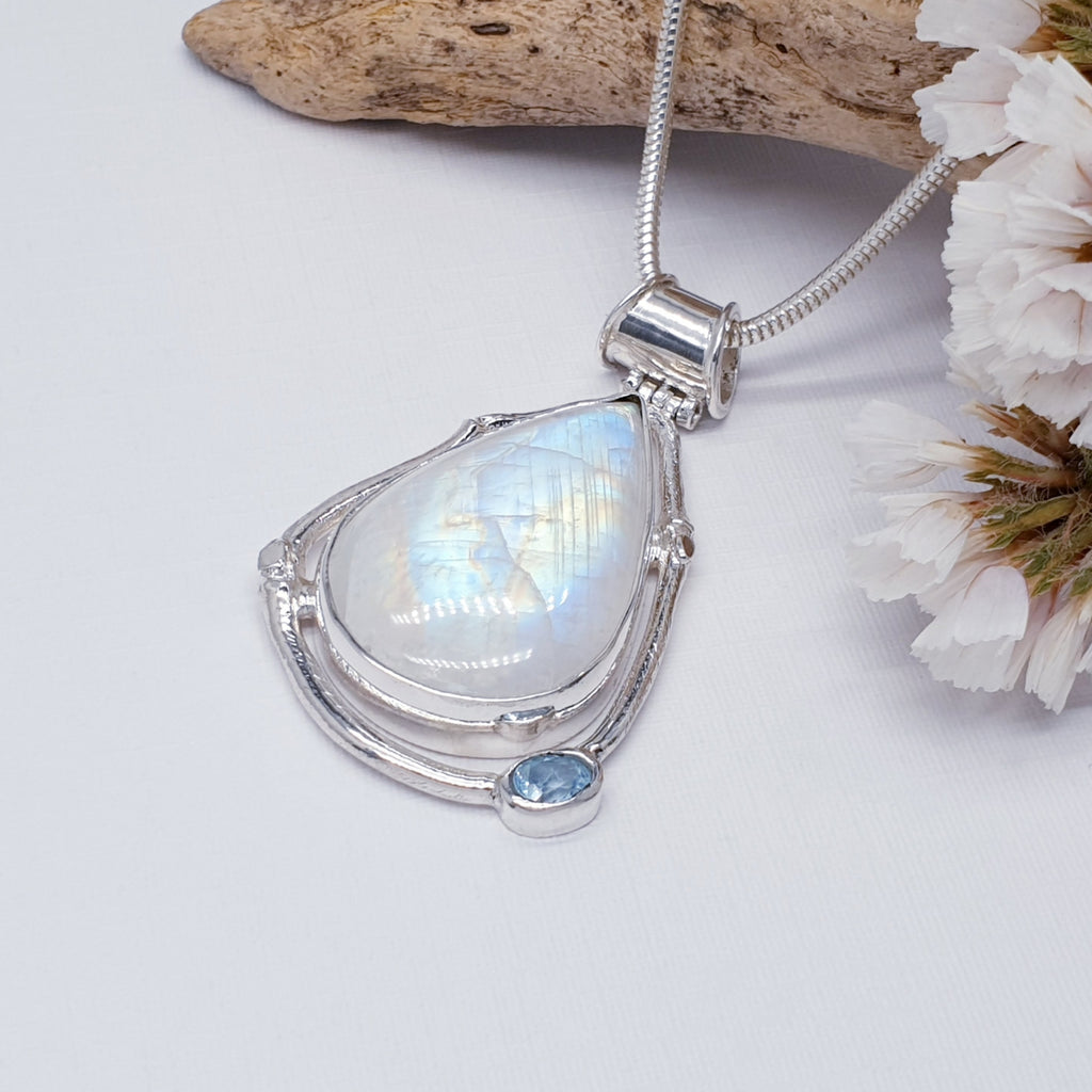 Our one of a kind Moonstone and Blue Topaz Aditi pendant on a heavy snake chain, photographed against a white background, with wood and white flowers as decoration