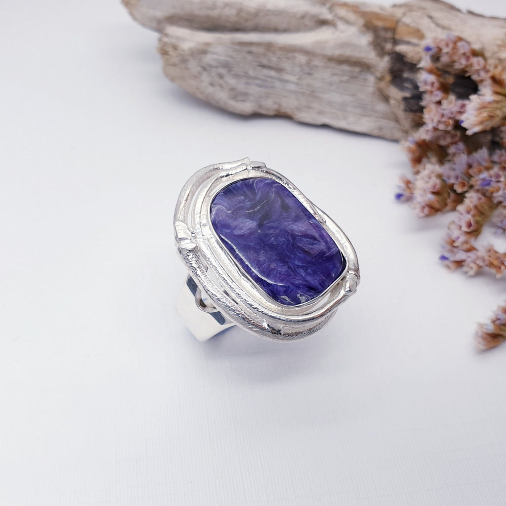 Charoite ring on a white background with bark and pink and white foliage to the right of it