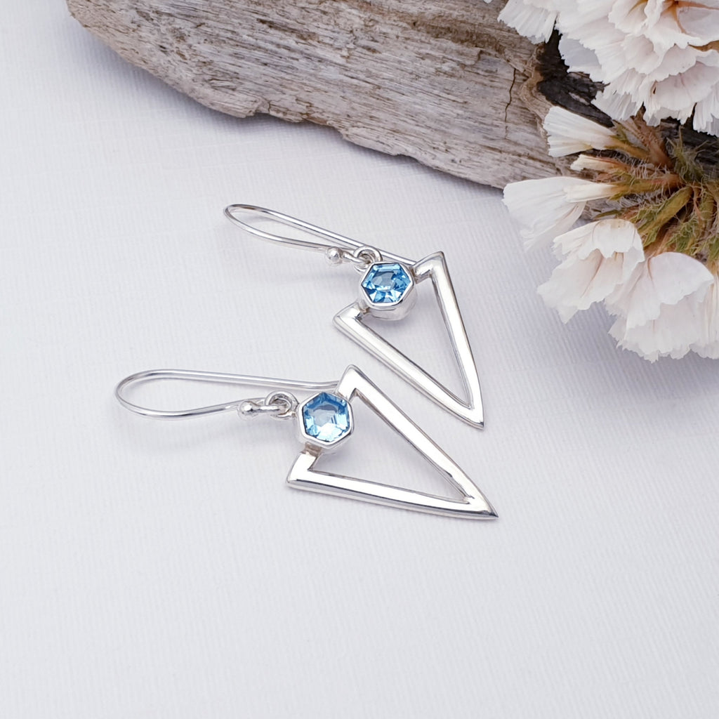 Blue Topaz Acute Triangle earrings, with hooks. They are presented on a white background with bark and foliage.