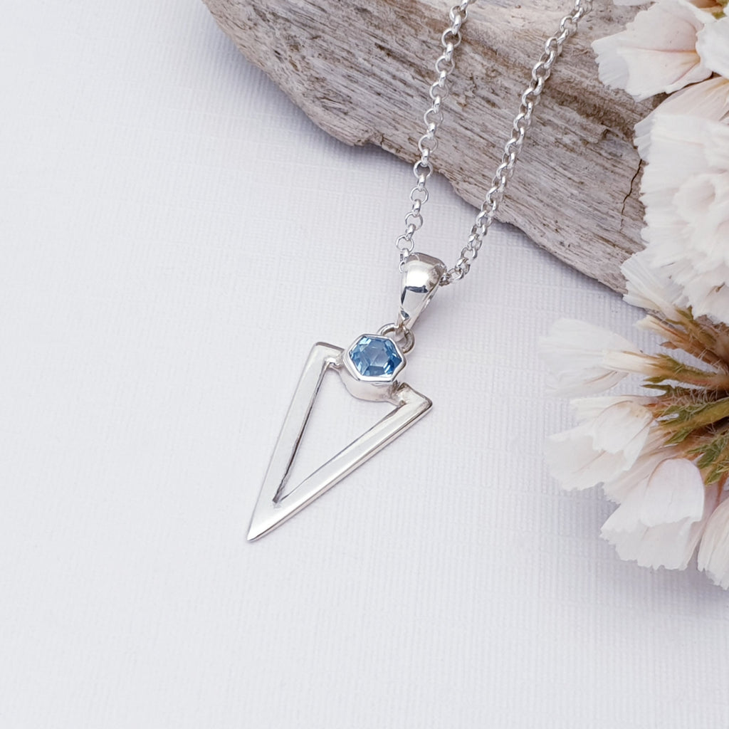 Blue Topaz Acute Triangle pendant on a medium belcher chain. On a white background with wood and foliage in the background