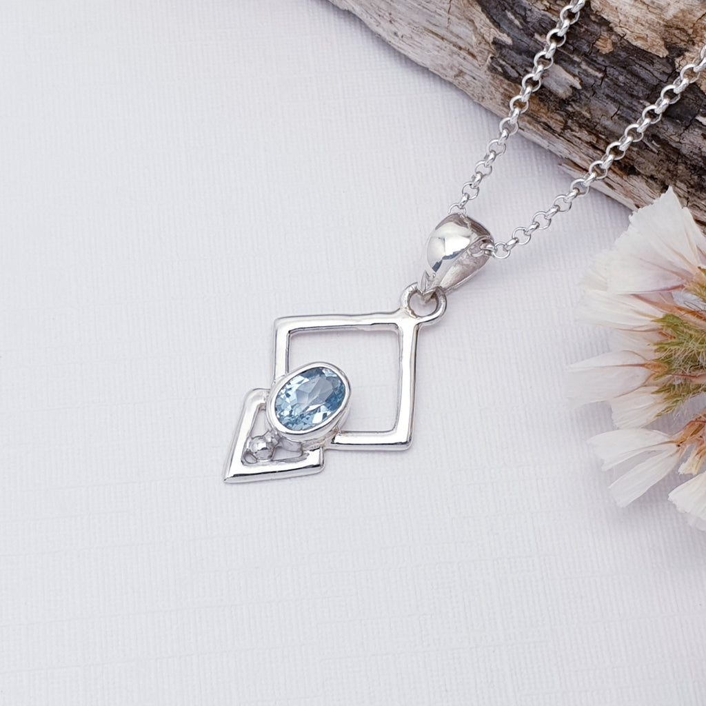 Blue Topaz Sterling Silver Triangulum Pendant on a white background with bark and white foliage on the top right of the image