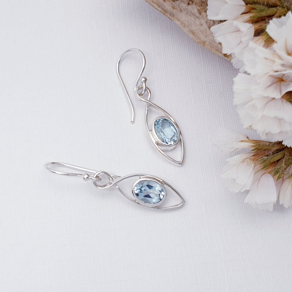 Blue topaz sterling silver raindrop earrings on a white background with wood and white foliage in the top right corner