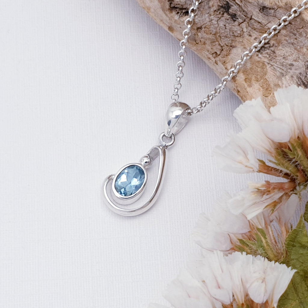 Blue topaz sterling silver midas pendant on a white background with bark and white foliage in the top right corner on the image