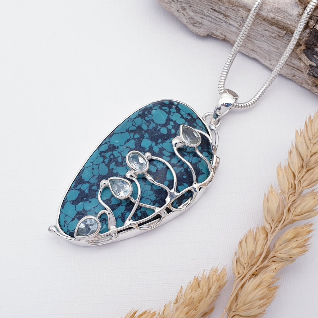 Our large one of a kind turquoise and blue topaz demeter pendant strung on a heavy snake chain, photographed against a white background with wood and autumn foliage as decorations.